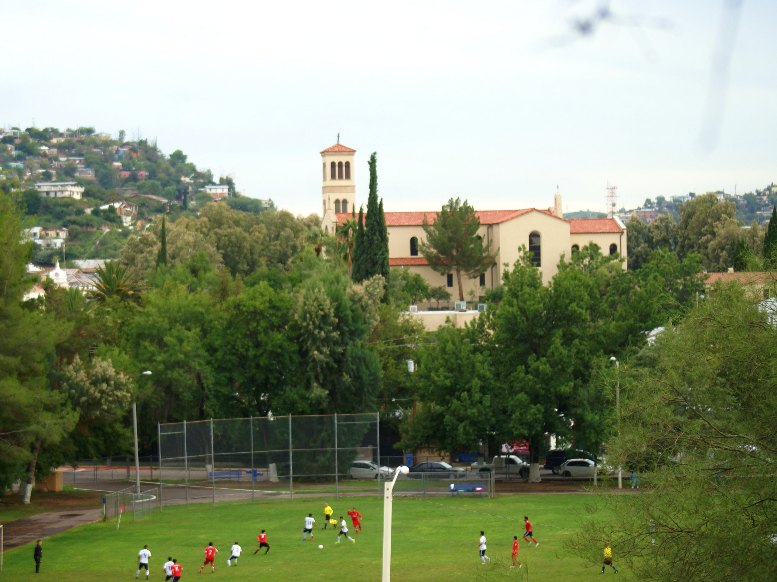 Beige city hall building stands in the background of a green soccer field where players in red and yellow run