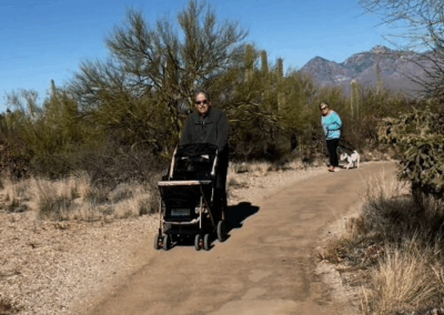 Image shows two hikers with a stroller and a leashed dog along a flat dirt road.