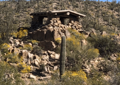 Image shows a rock cabin constructed by the Civilian Conservation Corp among a forest of shrubs and saguaro cacti.