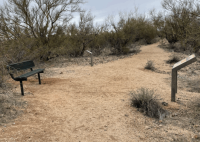 Image shows a flat, wide trail between a bench and several interpretive signs along the hike.