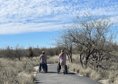 Image shows hikers using wheelchairs strolling side-by-side along the wide paved trails.