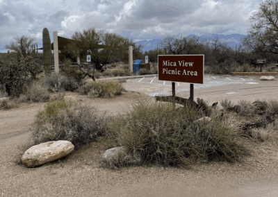 Image shows a sign that designates the Mica View picnic area, with a shade ramada and picnic tables nearby.
